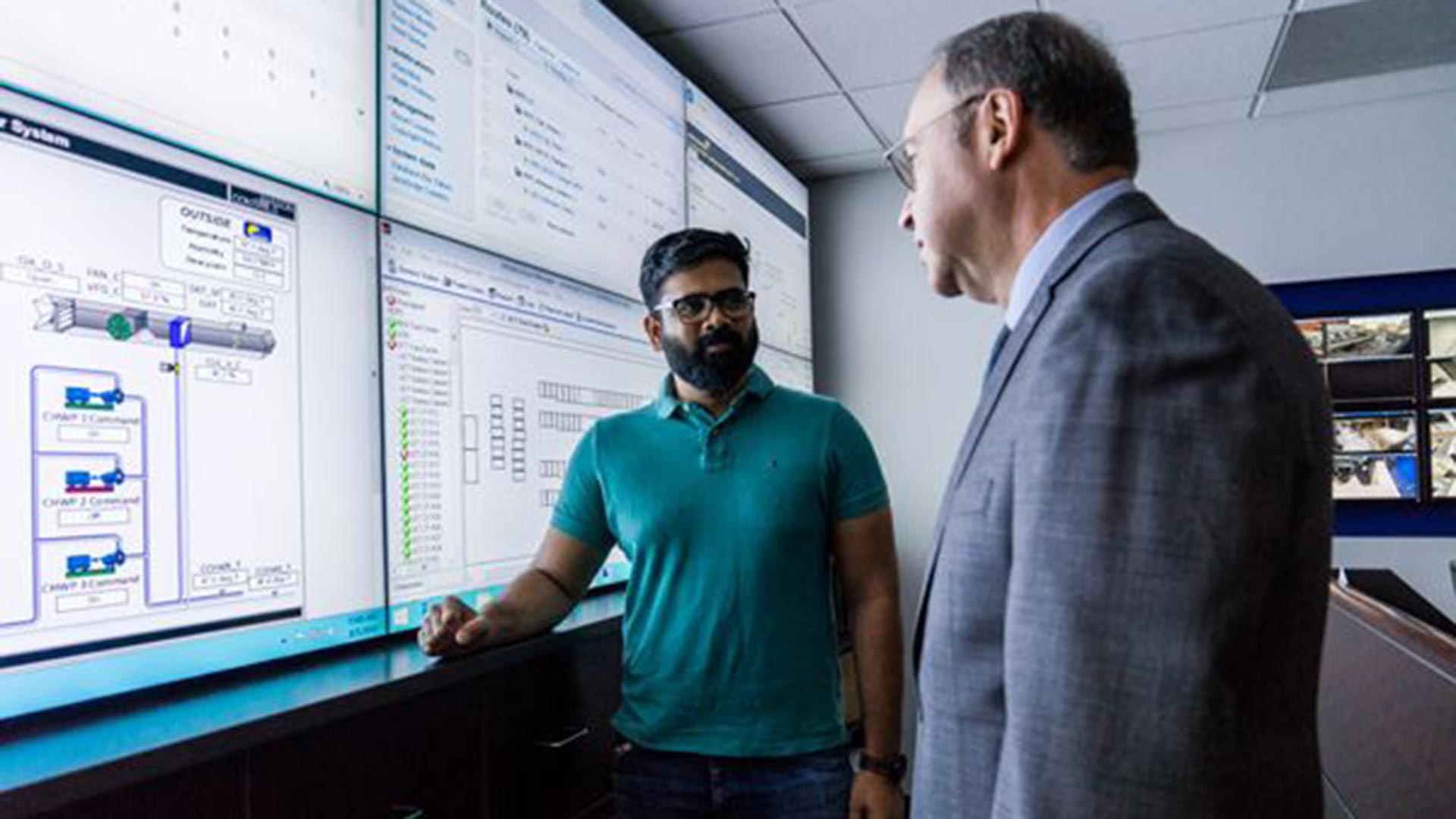A young technician in a teal polo and glasses stands in a control room beside a senior professional in a gray suit, pointing to multiple large wall-mounted monitors displaying technical diagrams and data.