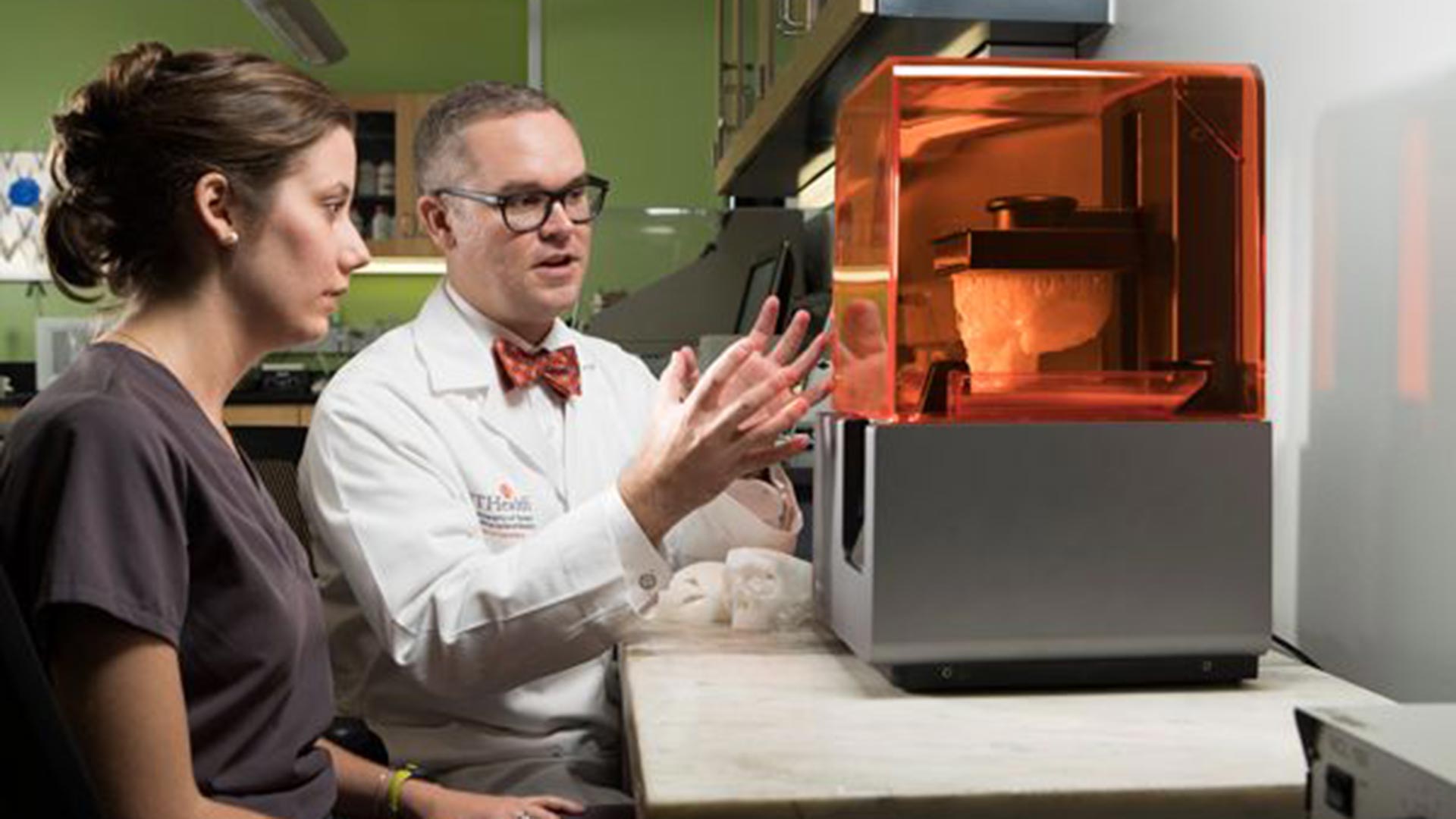 A female researcher in gray scrubs and a male faculty member in a white lab coat and orange bow tie seated beside a small 3D printer with an orange-tinted cover, discussing the anatomical model printing inside.