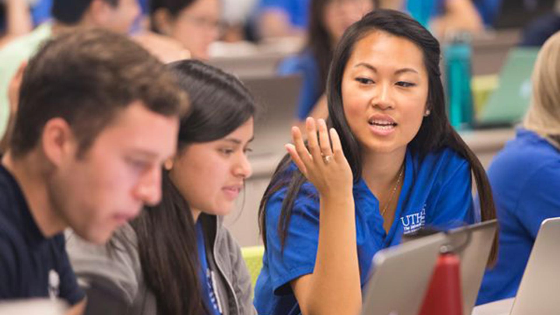 A young woman in a blue UTHealth Houston polo animatedly speaking and gesturing with her hand while seated at a table with two peers and their open laptops in a busy lecture hall.