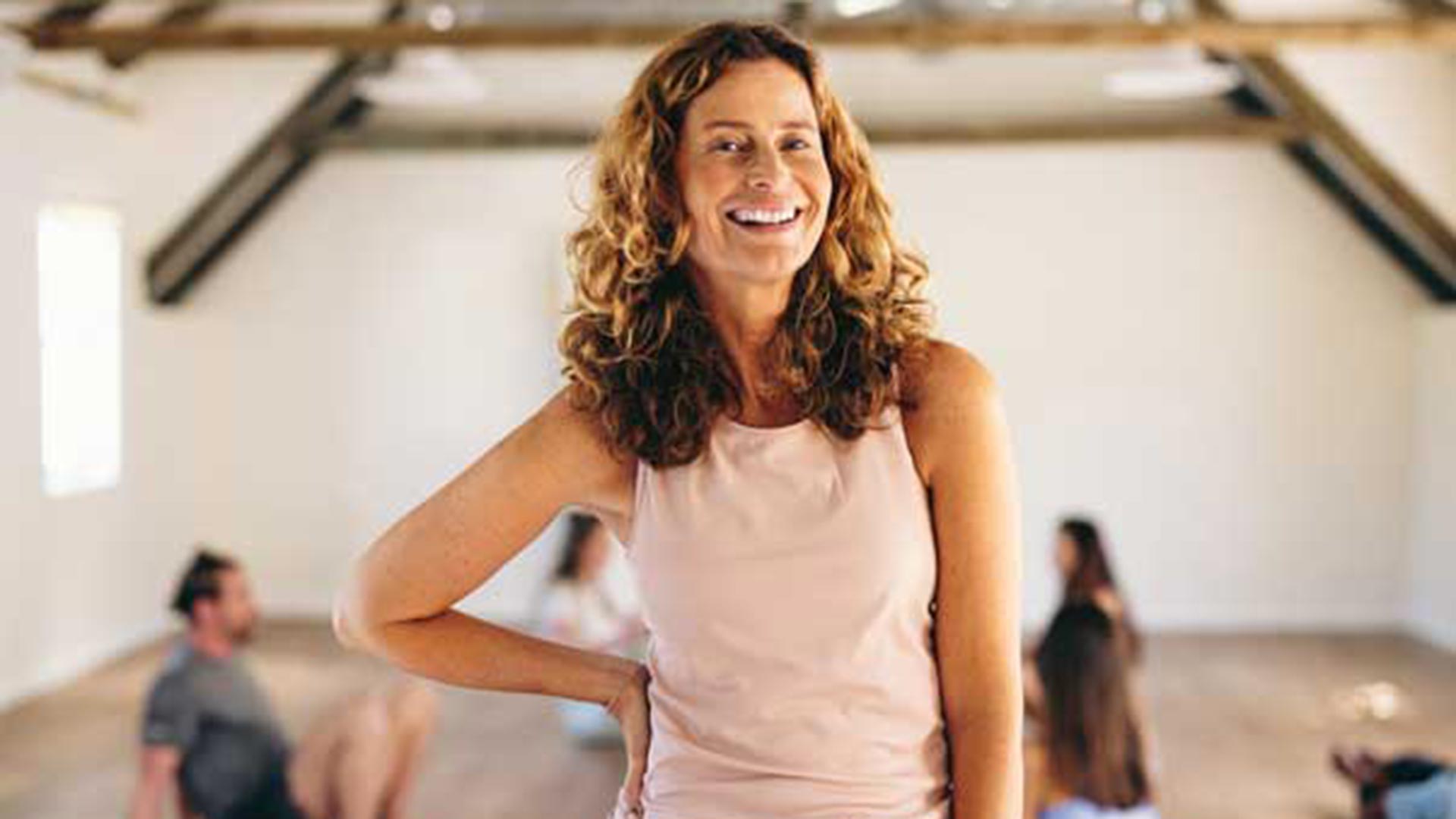 Smiling middle-aged woman in a pale pink sleeveless top standing confidently with one hand on her hip in a bright exercise studio, with blurred participants seated on mats behind her.
