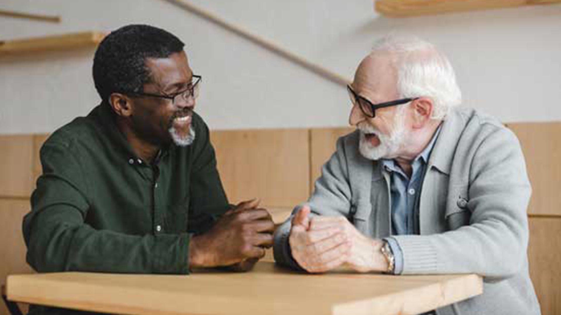 Two older men—one African American in a dark green button-up shirt and glasses, the other Caucasian with white hair and beard wearing a light gray cardigan—sitting at a wooden table, smiling and engaged in animated conversation in a bright room.