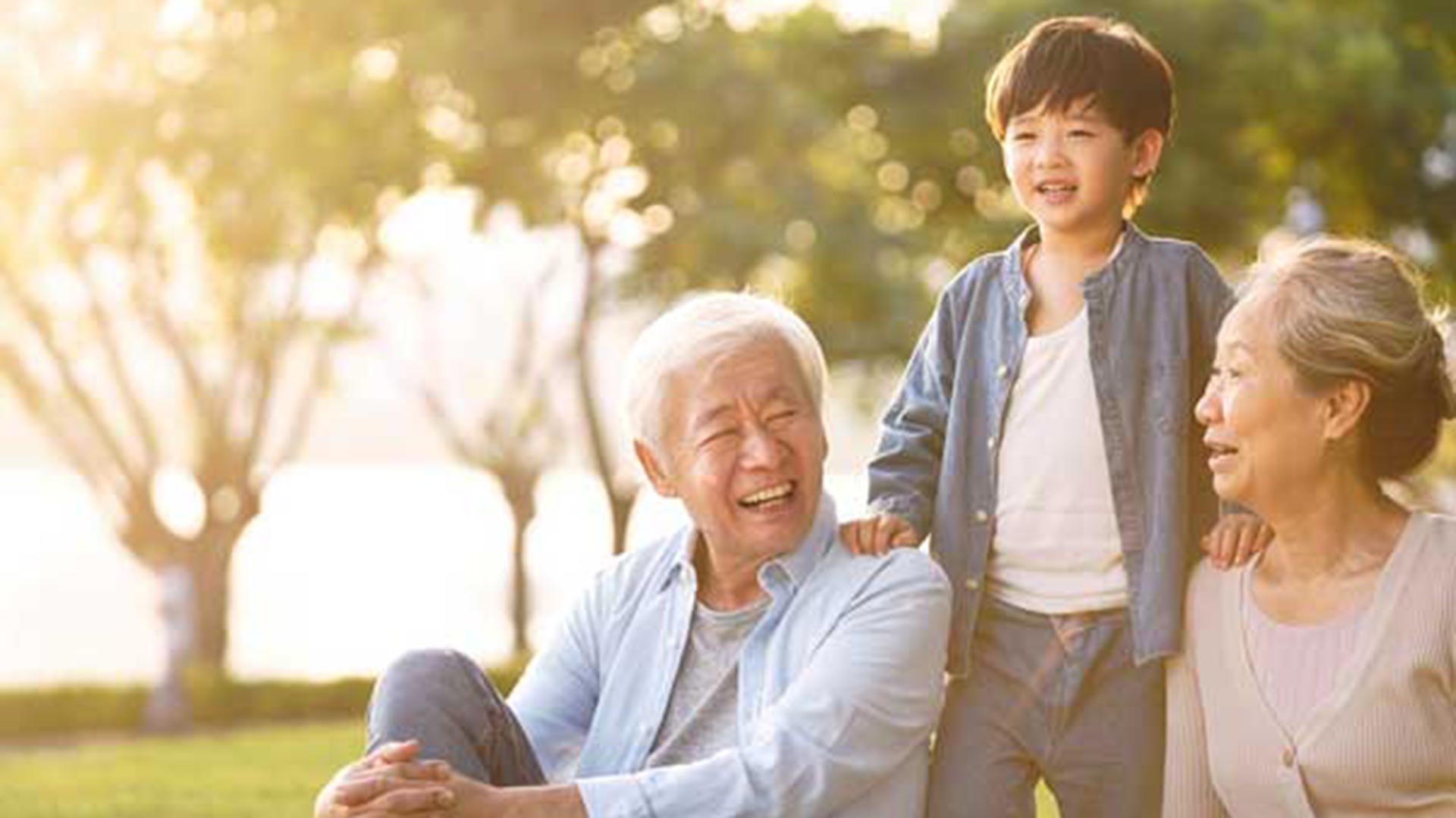 Elderly Asian man and woman sitting on a sunlit grassy area by the water, smiling warmly as a young boy stands behind them with his hands resting on their shoulders.