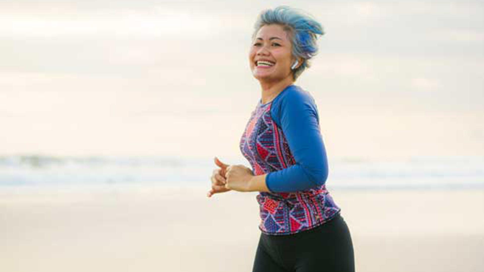 Mature woman with short blue-gray hair in a patterned long-sleeve running top and black leggings, jogging along a sandy beach with earbuds and smiling broadly.