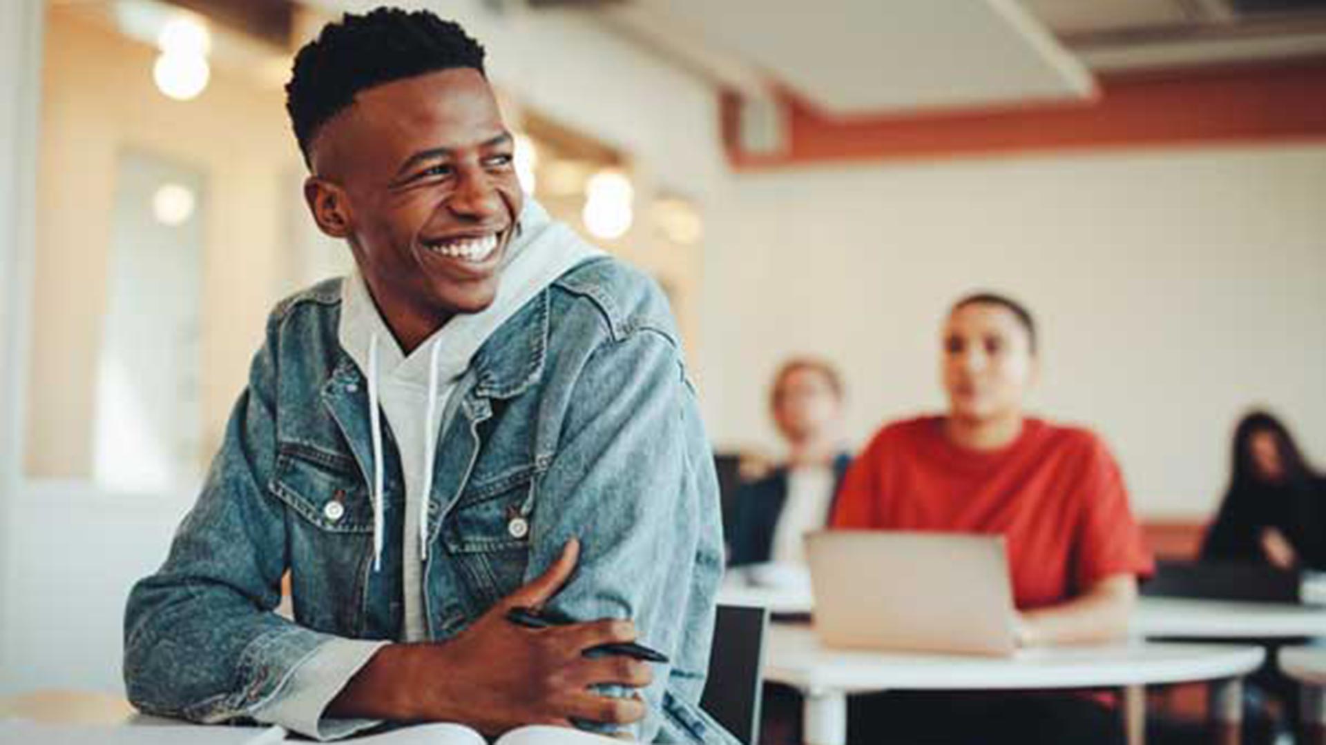 A young male student in a denim jacket layered over a white hoodie, seated at a bright classroom table, smiling and looking to his left with blurred classmates working on laptops behind him.