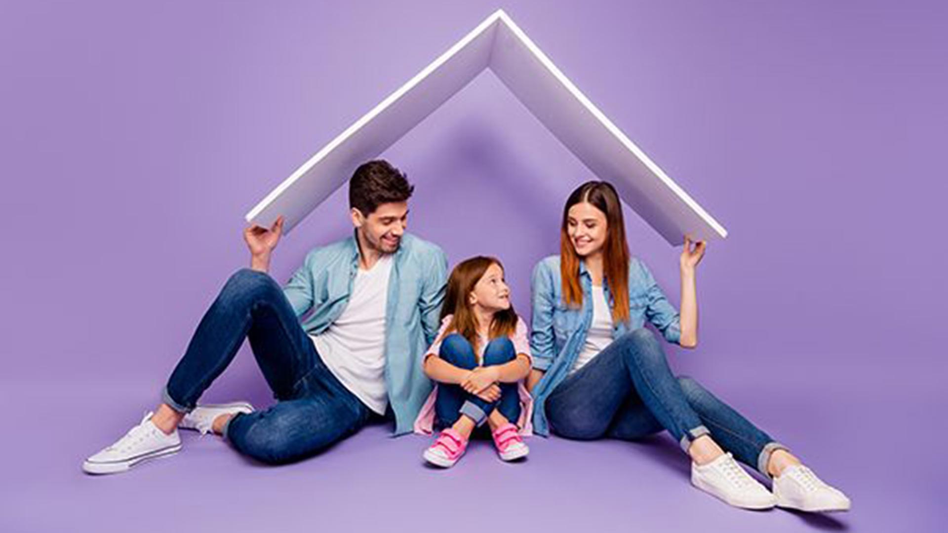 A mother, father, and young daughter seated on a purple floor, smiling as they hold a white panel overhead in the shape of a house roof.
