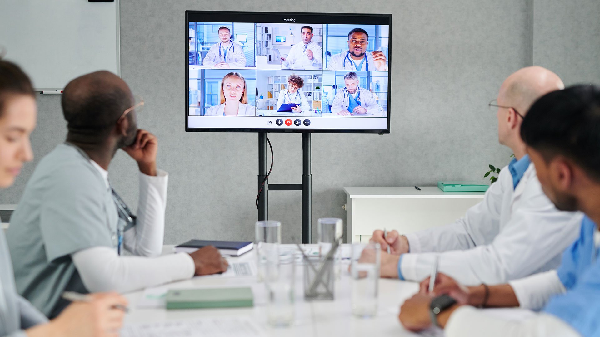 A group of medical professionals sit around a conference table watching a large screen displaying a virtual meeting with six doctors in separate video call windows.