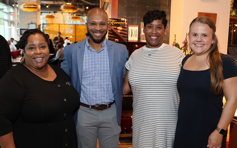 Four smiling attendees stand together for a group photo in a warmly lit restaurant, dressed in business-casual attire.