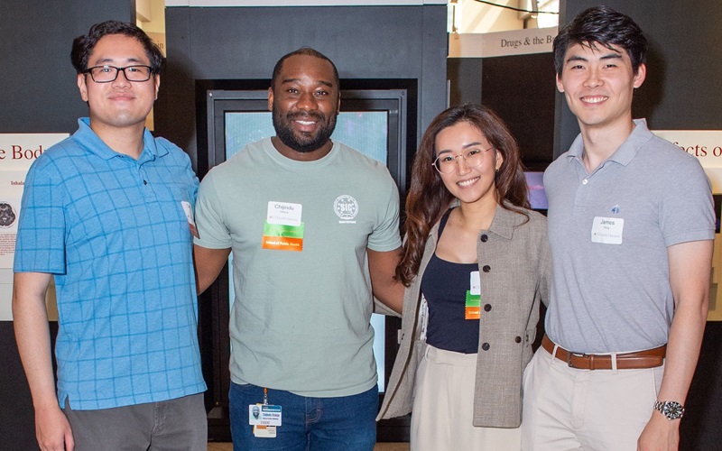 Group of younger alumni, one female and three male standing together and smiling for a photo