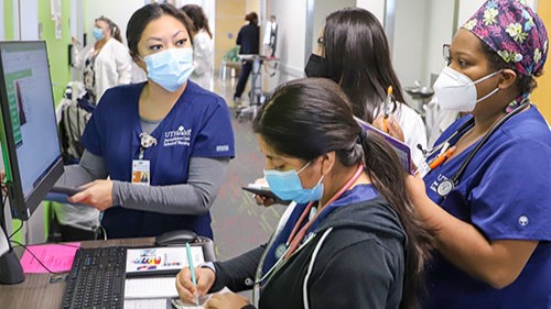 Masked nursing team at UTHealth Houston reviews information at a workstation while a trainee takes notes in a hospital hallway.