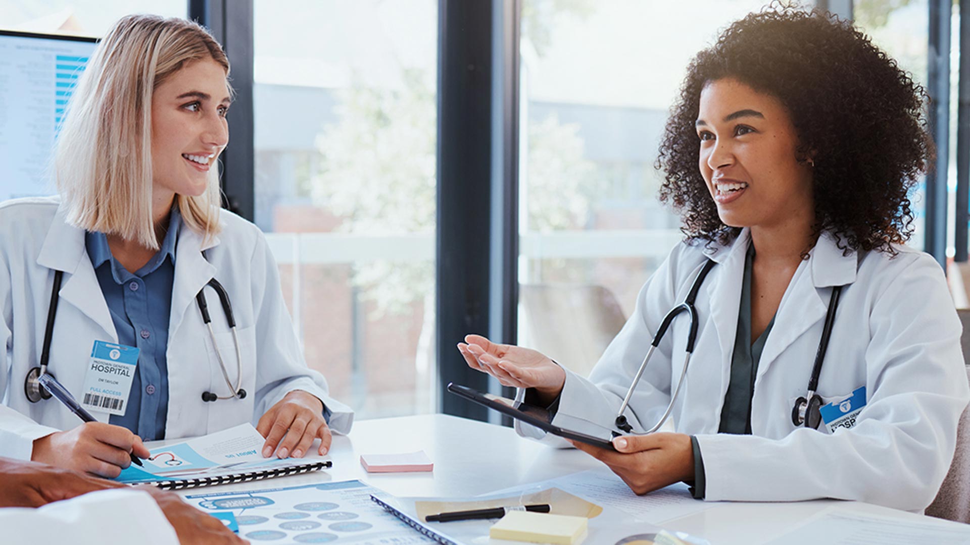 Two female doctors in white coats are seated at a busy conference table in a conference room with large computer monitors and wide glass windows. They are posed as if casually chatting with each other.