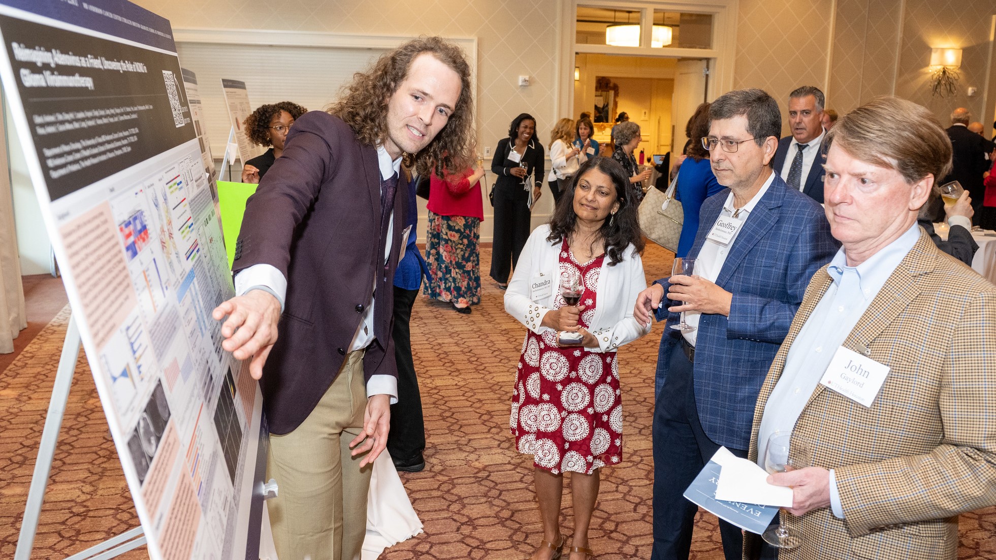 Researcher presents a scientific poster to three attendees at a reception, pointing to charts while guests with name badges listen and hold drinks.
