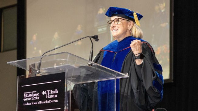 Smiling commencement speaker in blue-and-black academic regalia at a podium with MD Anderson and UTHealth Houston Graduate School of Biomedical Sciences signage.