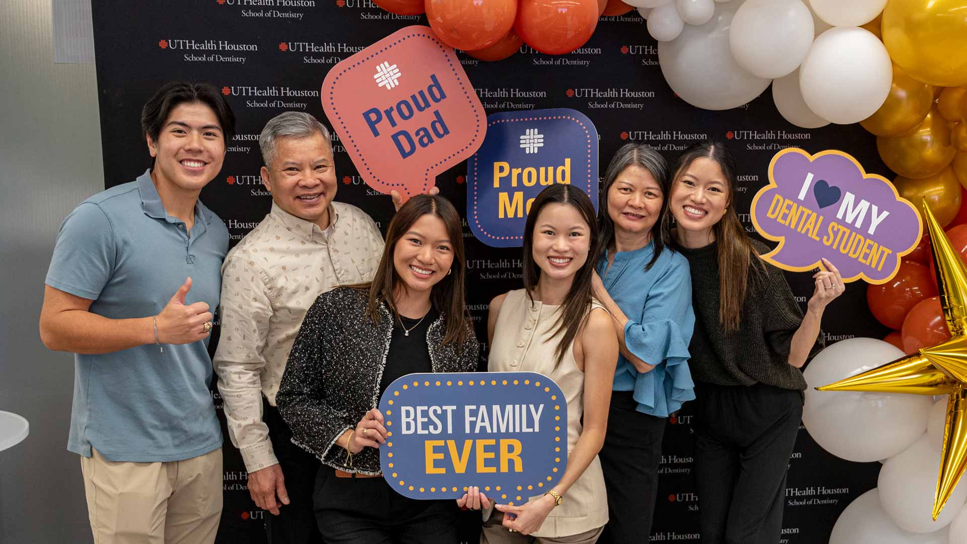 Family of six smiling and posing with signs such as 'Best family ever' and 'I love my dental student' during a community event.