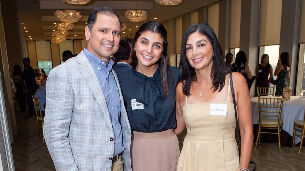 Smiling trio—two adults and a young woman—posing together at a reception in a bright banquet hall with round tables and gold chairs, wearing name tags.