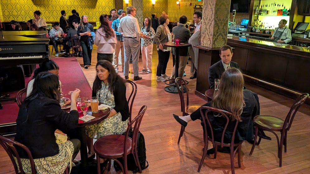 Reception in a lounge with wood floors and warm lighting; guests chat at small round tables and cocktail tables near a bar and a grand piano.
