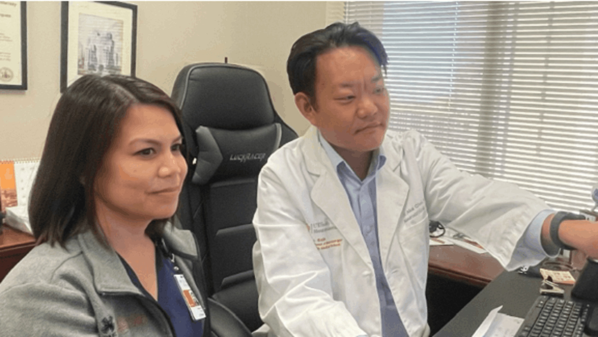 Two UTHealth Houston medical professionals, one in a white coat and the other in scrubs, sit side-by-side at a desk in a clinical office, looking attentively at a computer screen during a collaborative work session.