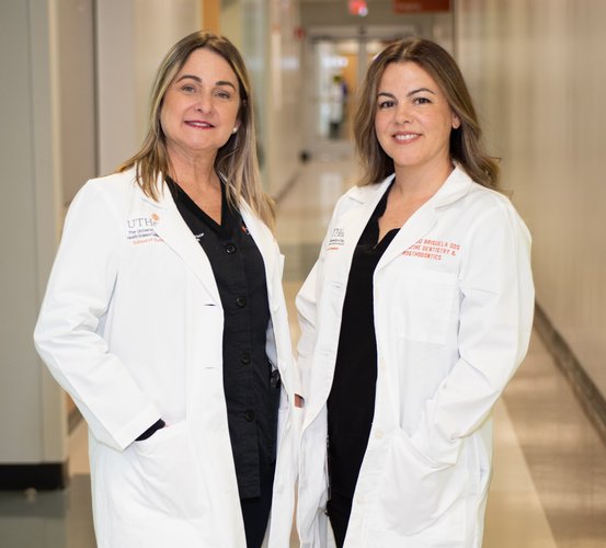 Maria A. Loza, DMD, MS (left), and Claudia Ruiz-Brisuela, DDS (right), stand with their hands in their white coat pockets in a clinical hallway at UTHealth Houston School of Dentistry.