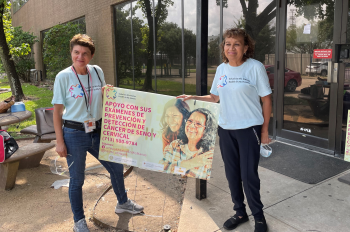 Two women hold a sign outside of an event informing hispanic women about cervical and breast cancer screenings.
