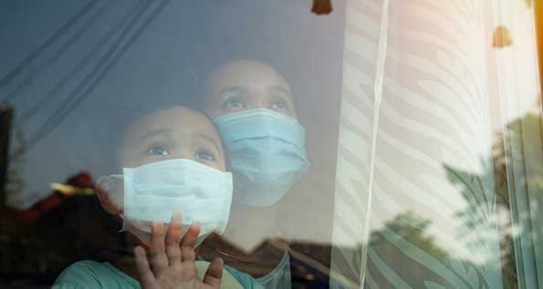 Image of a mother and daughter wearing surgical masks and looking out of a window. The child is touching the glass.