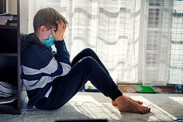 Photo of boy wearing mask with head in hands. Mental health-related visits to hospitals for children and teens has increased since the COVID-19 pandemic began due to increased stress. (Photo courtesy of Getty Images)
