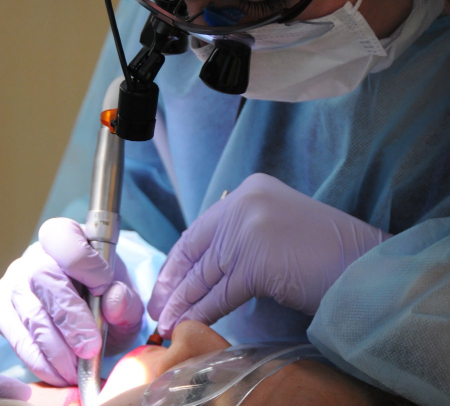 UTHealth student in masks, gloves and wearing loupes performs a dental procedure on a patient who is wearing plastic eye protection.