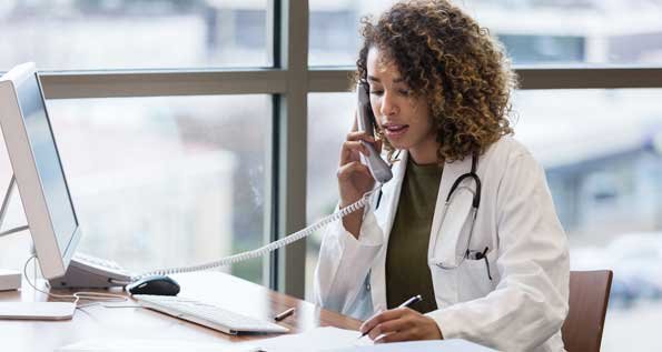 Photo of a woman wearing a doctor's white coat, taking on the phone at a desk with a computer. (Photo by Getty Images)