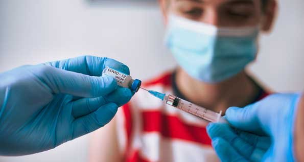 up-close photo of a gloved health care provider inserting a needle into a vial of vaccine, with a masked adolescent boy in the background. (Photo by Getty Images)