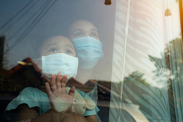 Photo of child looking out the window with a mask on. Physicians say getting to the bottom of why most children seem to be spared from severe illness related to COVID-19 could be the key to understand how to treat adults. (Photo credit: Getty Images)