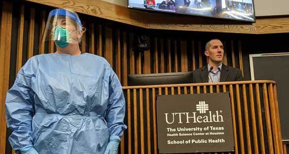 Image of Scott Patlovitch speaking at the school of public health. A person in personal protective gear needed to serve coronavirus patients sands to the left of him.