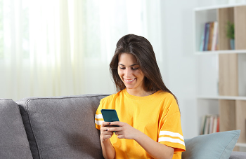 Photo of young woman on a sofa looking at her phone and smiling (Photo courtesy of Getty Images)