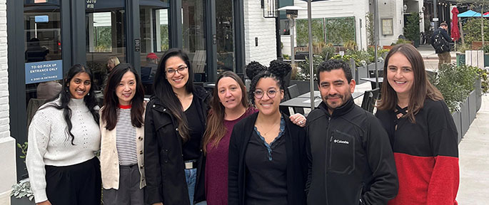 Seven members of a research lab stand together outdoors in front of a modern restaurant patio, smiling at the camera; casual winter clothing, glass storefront and planters in the background.