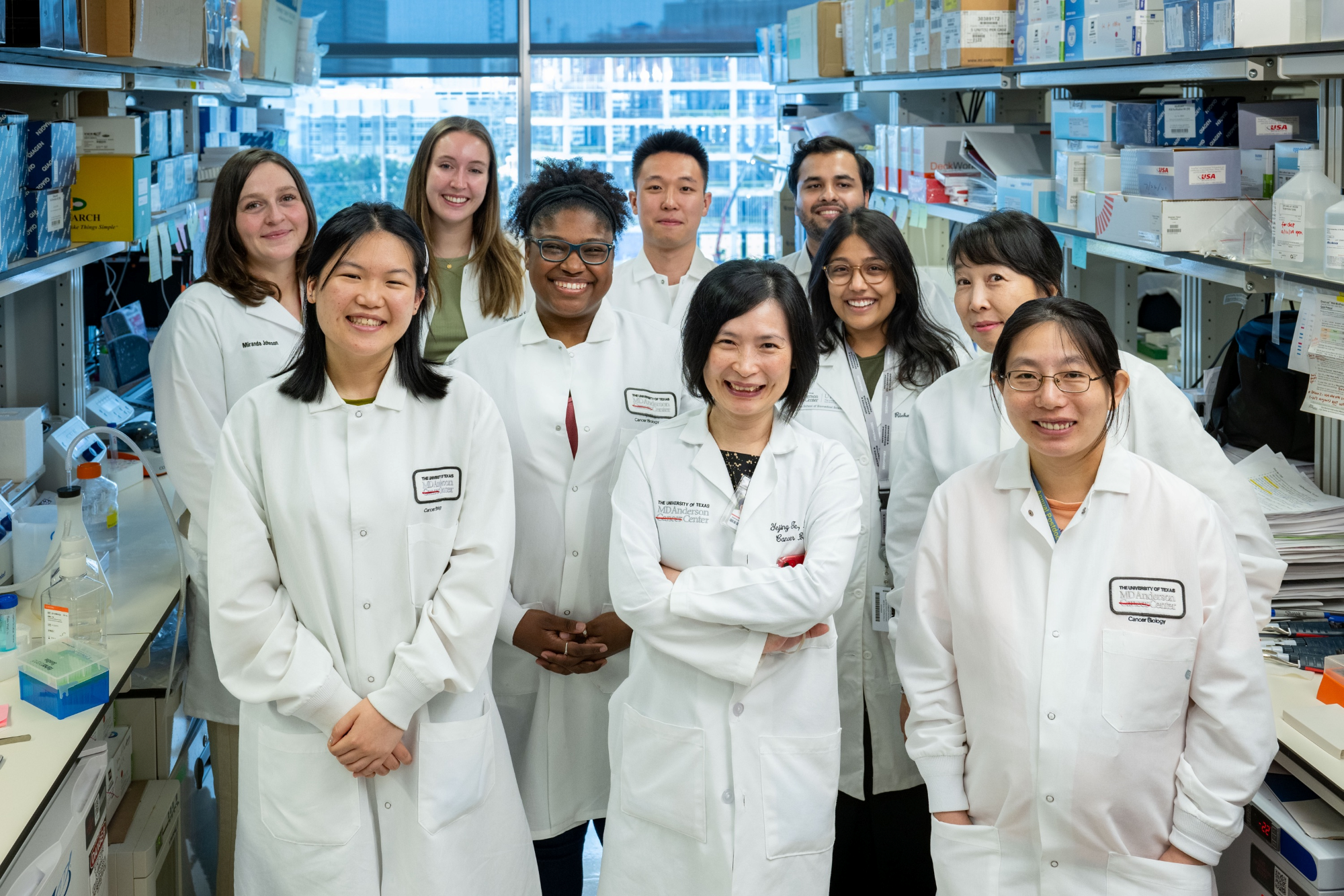 Research team of eleven scientists in white lab coats standing in a biomedical lab aisle, central PI with arms crossed, benches and equipment on both sides and a city skyline visible through the windows.