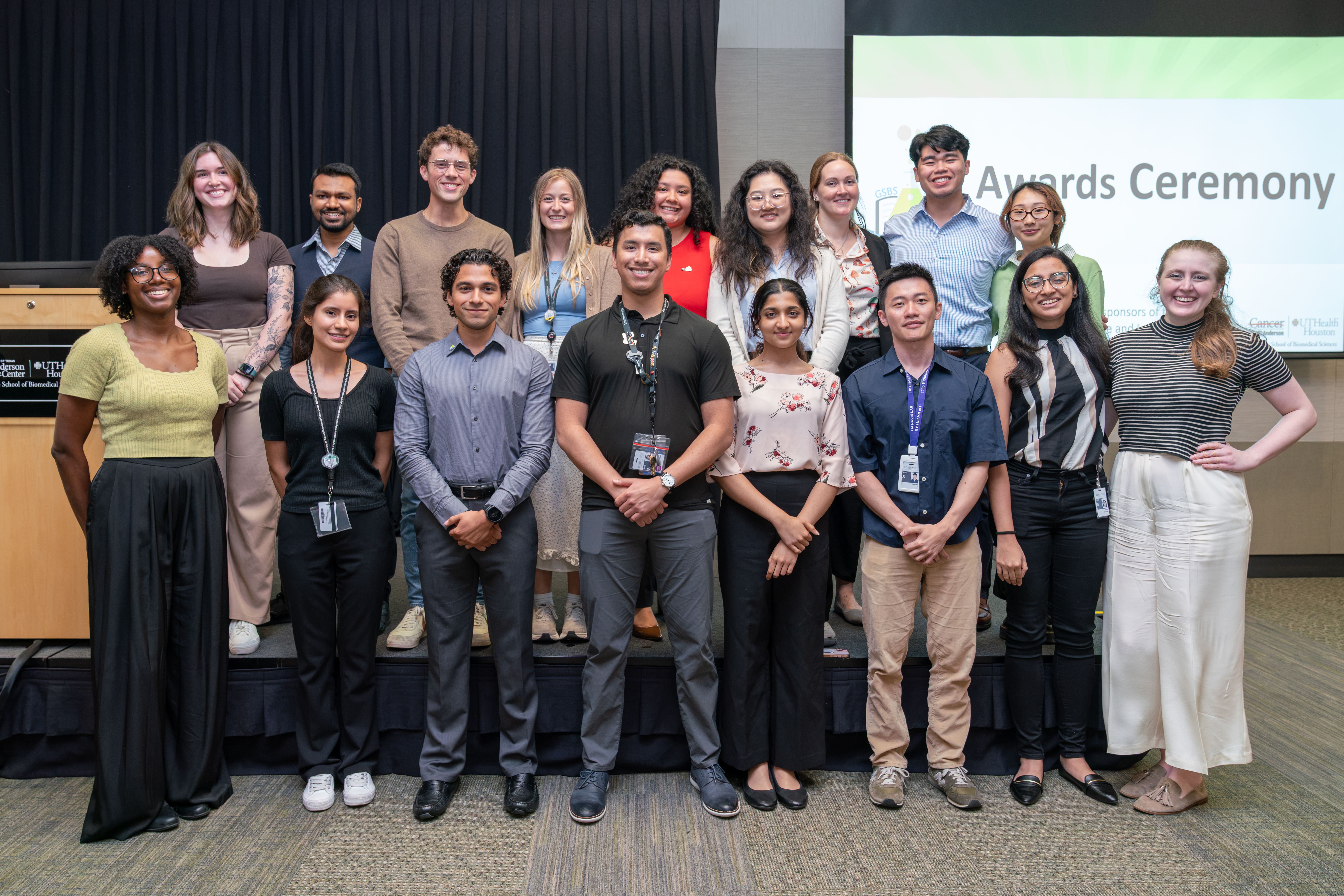 Group photo of students and staff posing on a stage at an awards ceremony with a presentation screen in the background