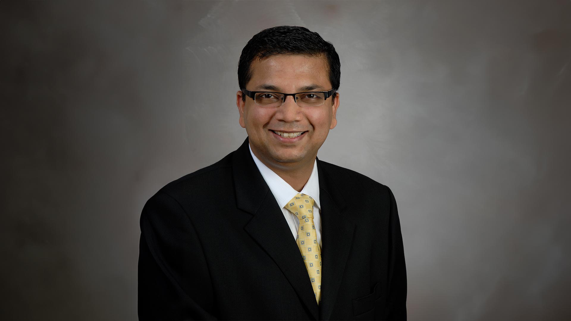 Dr. Nirav Thosani poses for a professional headshot in a black suit with yellow tie in front of a gray brackground.