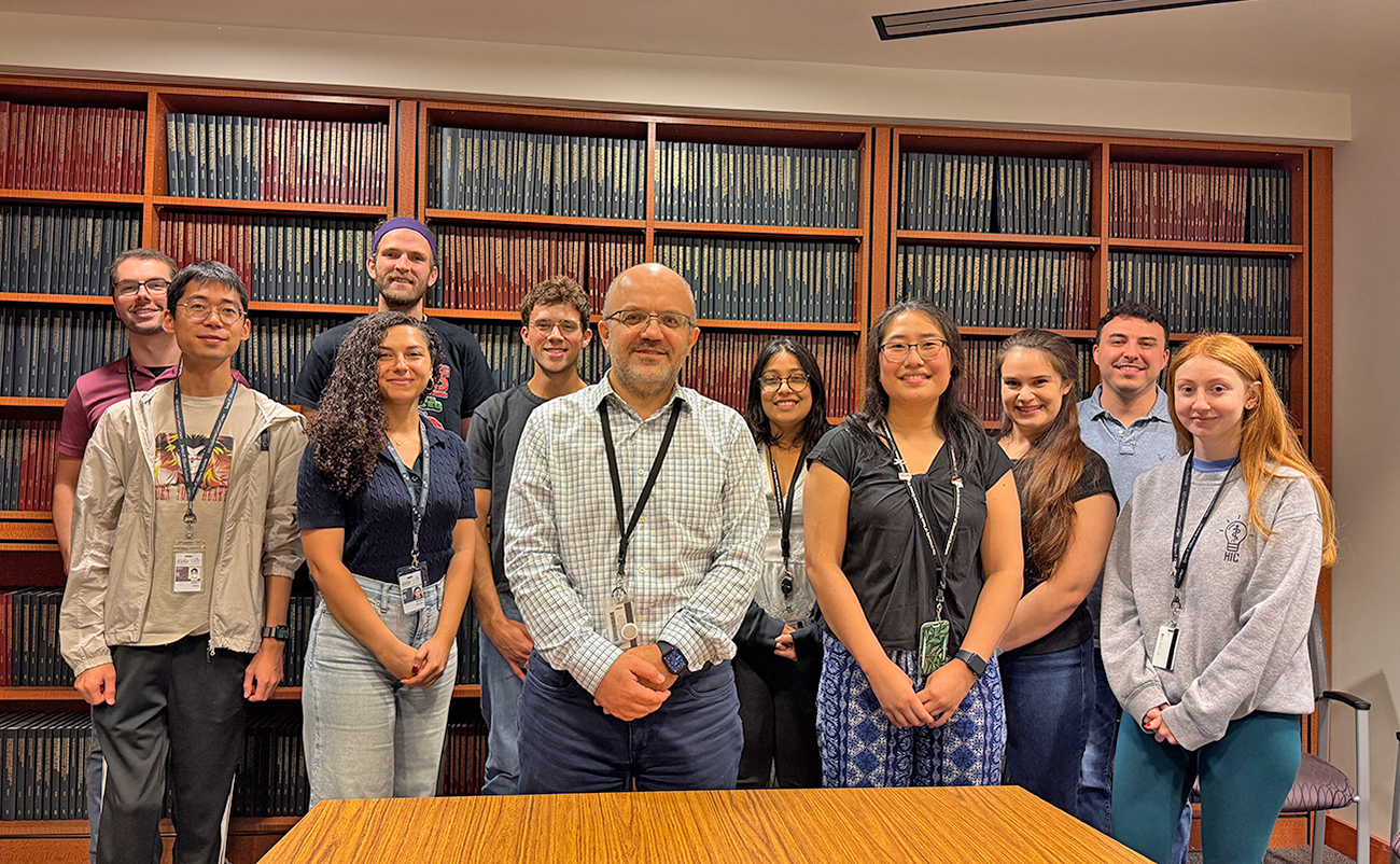 Group photo of eleven people standing in a library room, smiling in front of tall wooden bookshelves filled with bound journals; one person stands centered in front, others arranged in two rows around a conference table.