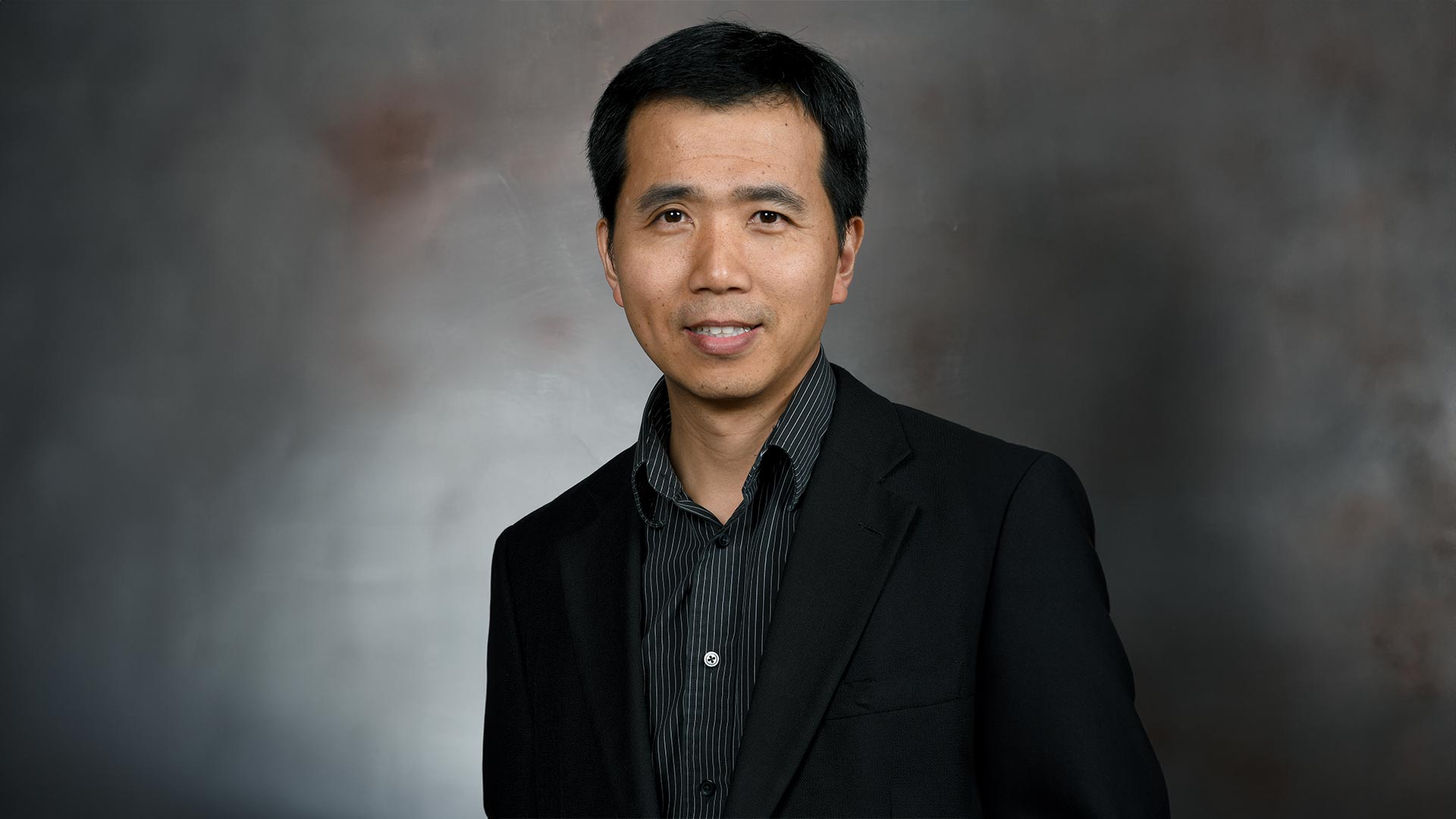 Dr. Qingchun Tong poses for a professional headshot in a black suite and black shirt in front of a gray background.