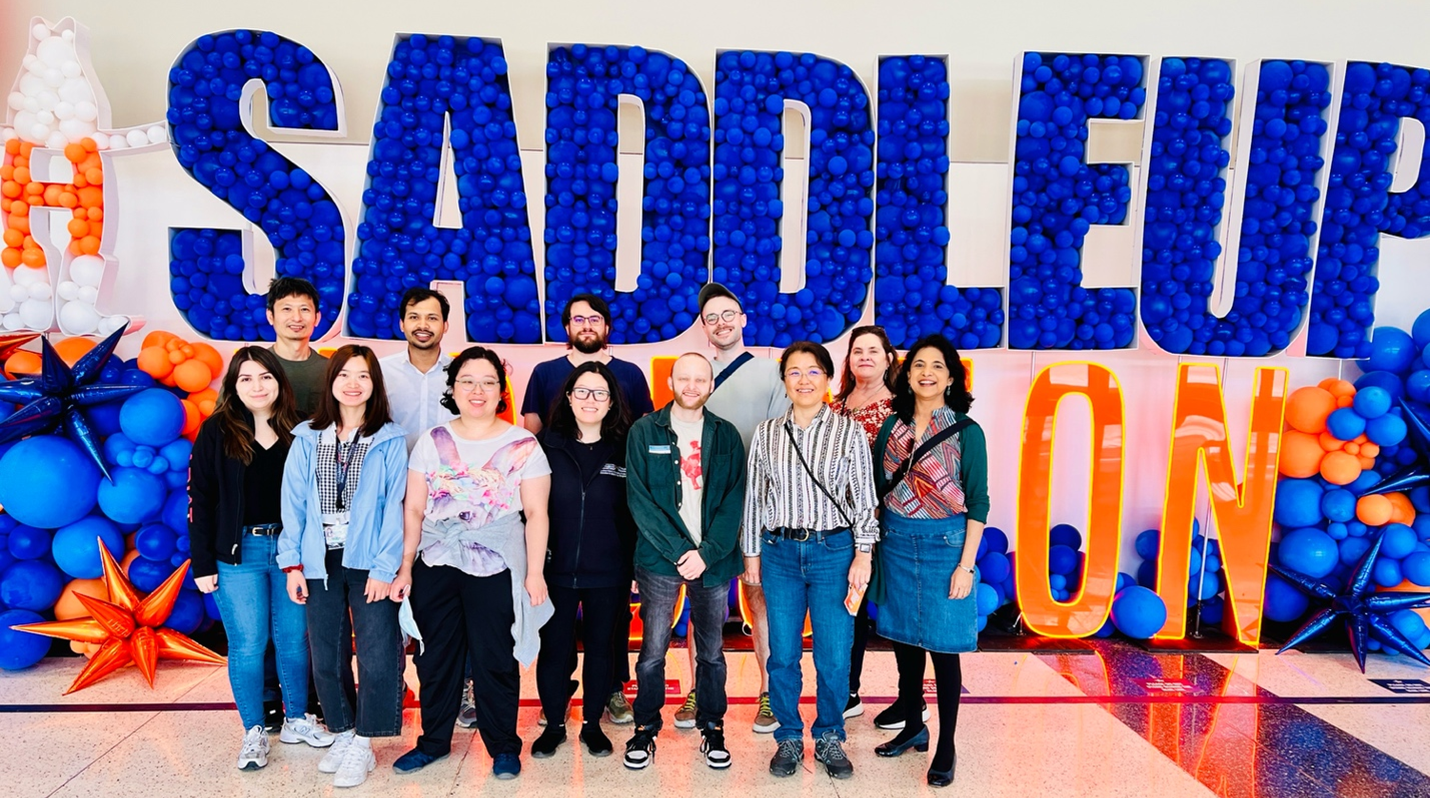 Group photo of the Arur Lab&mdash;twelve people smiling in front of giant blue and orange balloon letters spelling 'SADDLE UP ON' in a bright lobby.