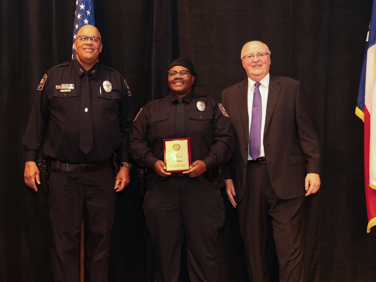 Two uniformed UT Police officers—center honoree holding a plaque—stand with a suited administrator onstage between U.S. and Texas flags.