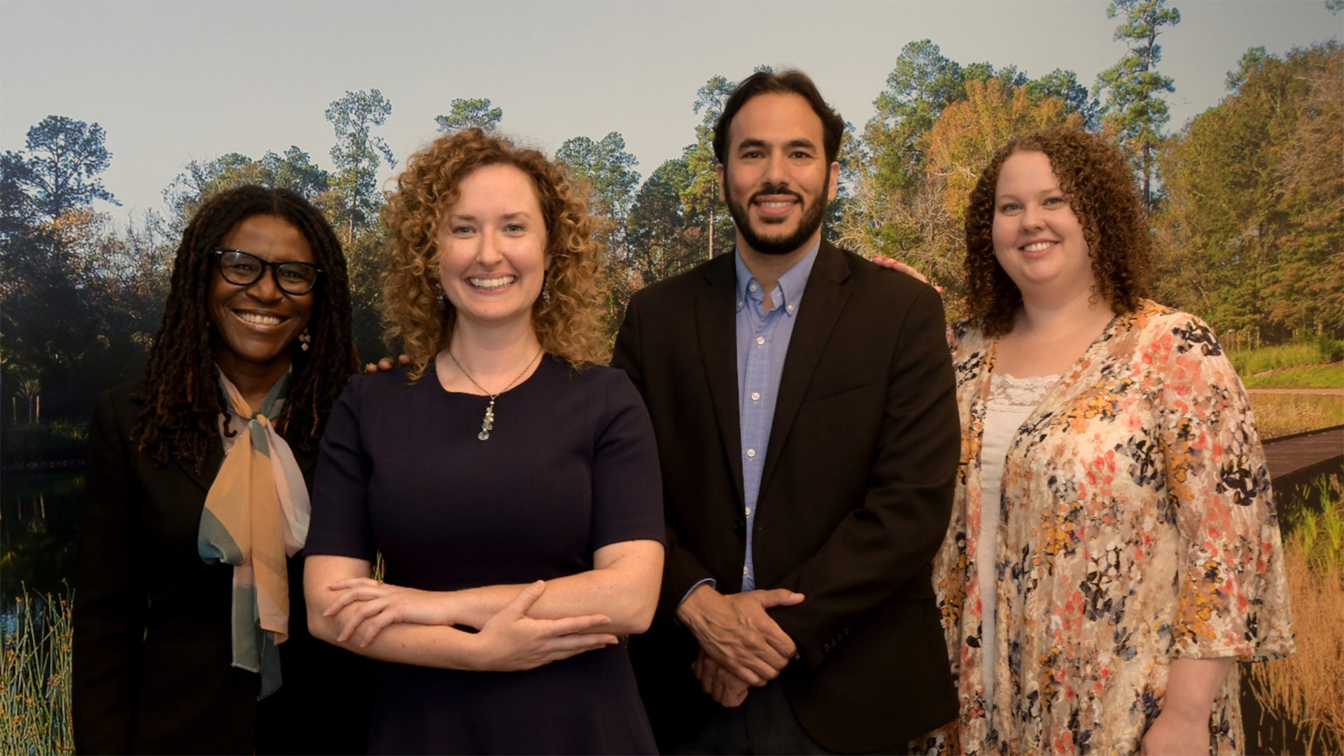 Four diverse professionals—a Black woman with glasses and a patterned scarf, a curly-haired woman in a navy dress, a bearded man in a dark blazer, and another curly-haired woman in a floral blouse—standing side by side and smiling in front of a scenic backdrop of trees and water.