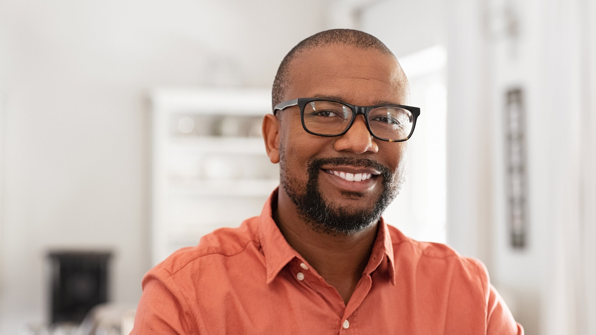 Smiling man wearing glasses and an orange shirt, standing indoors in a well-lit room with a blurred background.