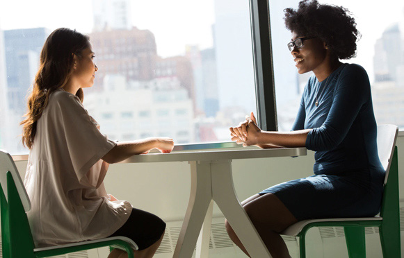 Two individuals sitting at a table in conversation, with city buildings visible through the large window behind them.