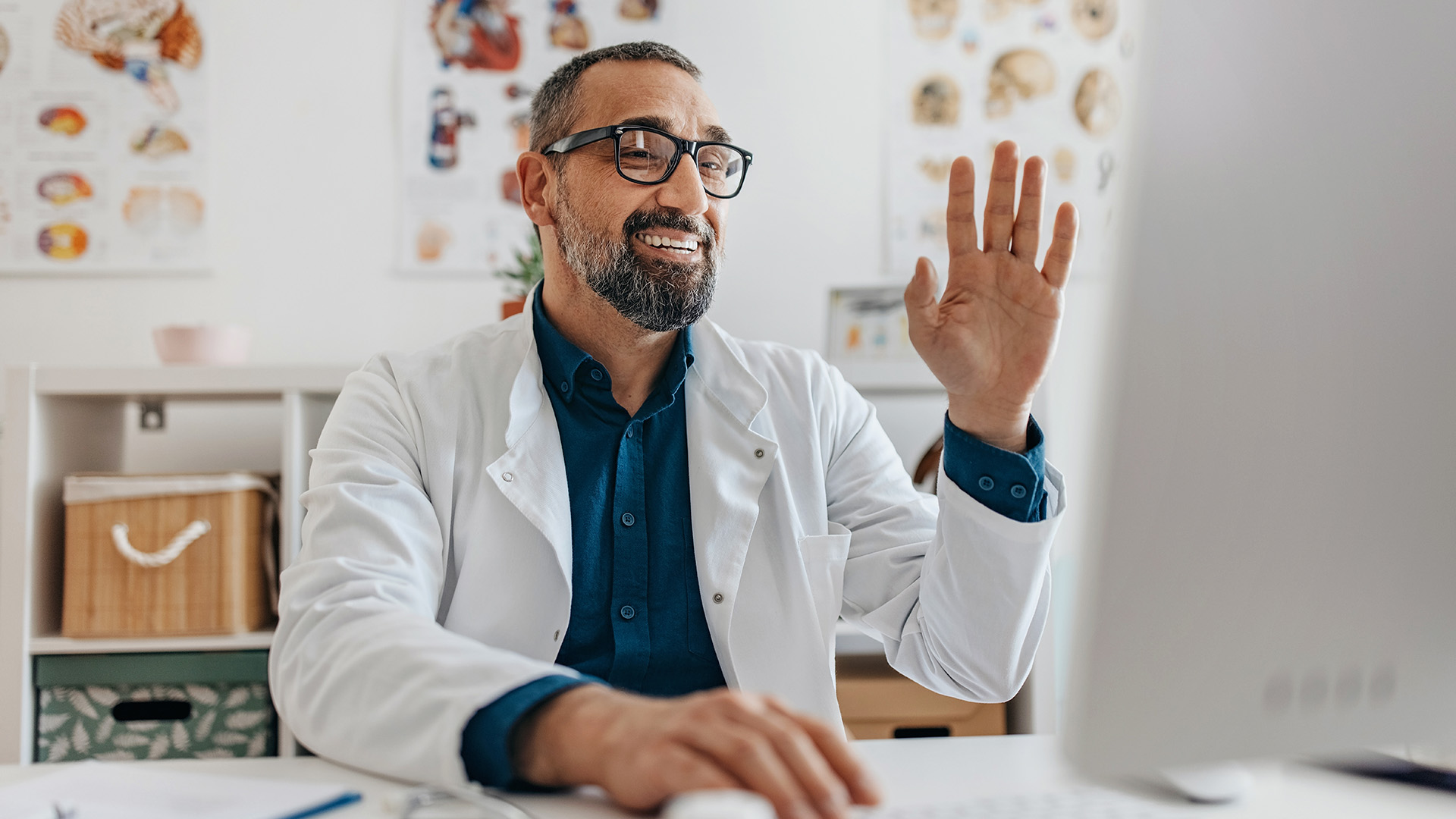 Smiling healthcare professional in a white coat waving during a video call in a medical office.