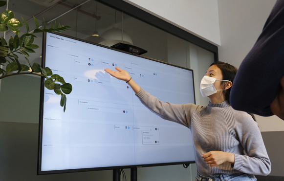 Individual wearing a face mask gestures to a large screen during a presentation, with another person observing.