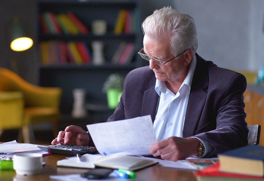 Older man sitting at a desk with papers a notebook, and a calculator. Courtesy Getty Images.