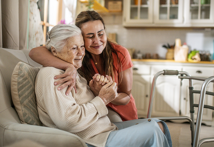 Younger woman hugging older woman on couch with walker nearby. Courtesy Getty Images.