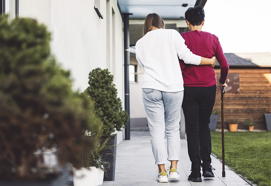 Younger woman helping older woman with cane walk. Courtesy Getty Images.