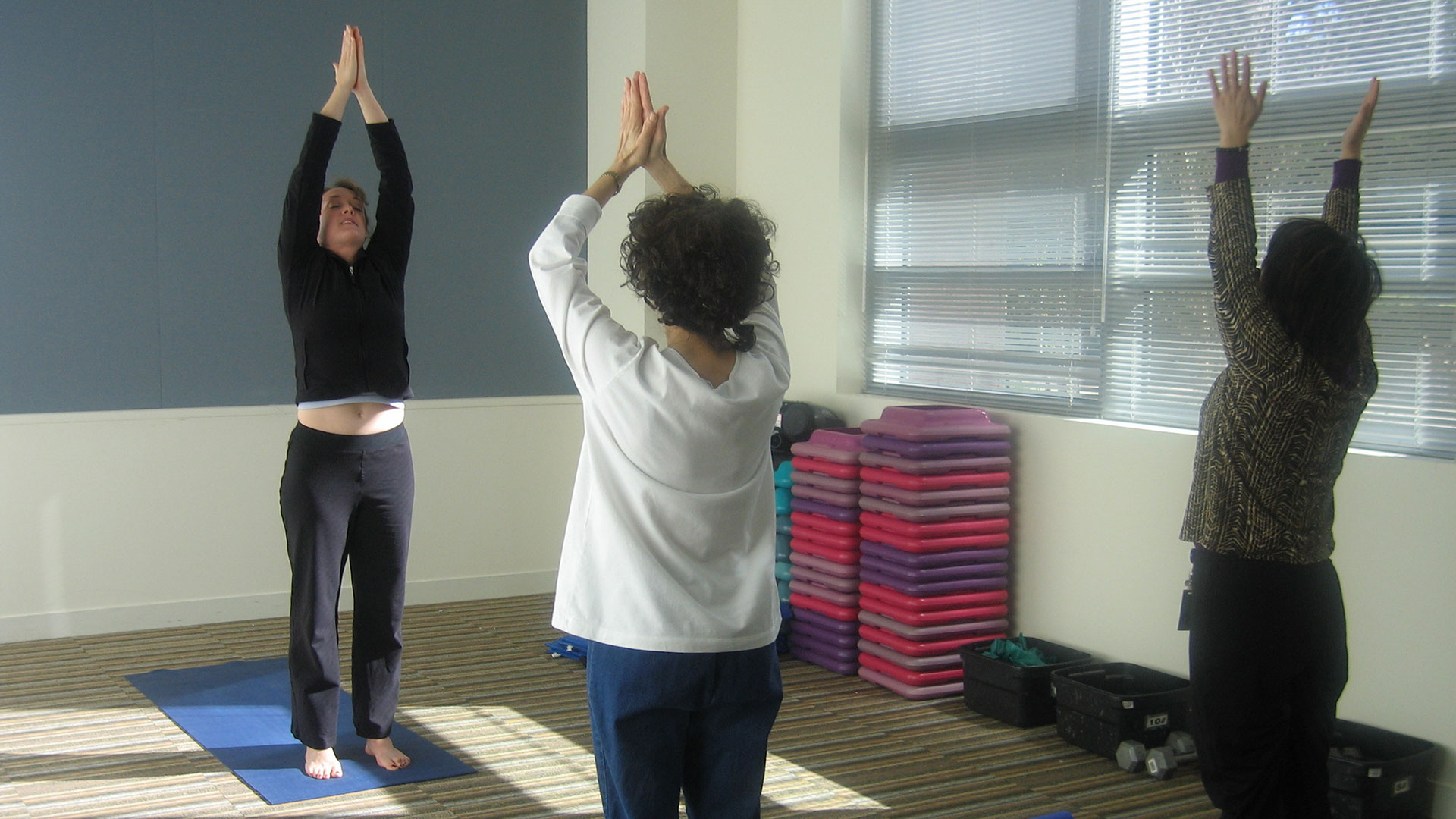 Three adults in a sunlit studio raise their arms overhead in a standing yoga pose, with stacked step platforms and weights near the windows.