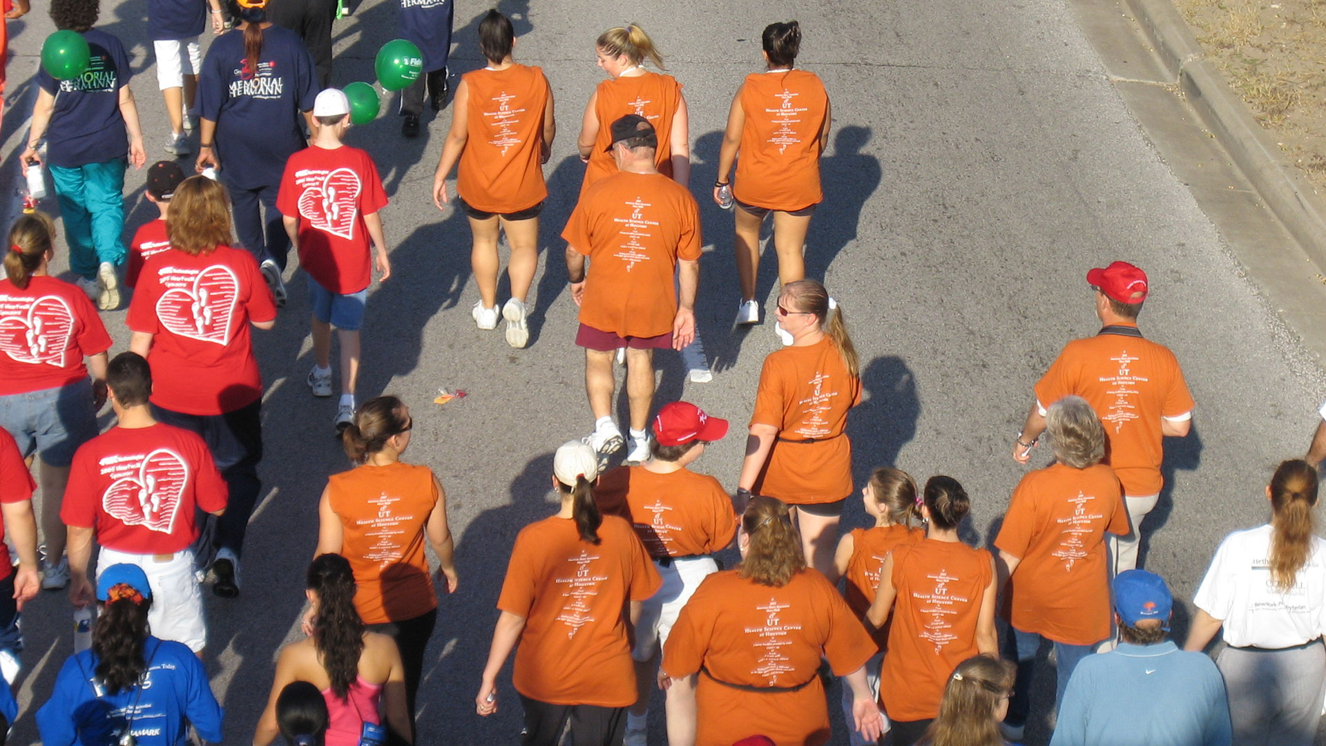 Overhead view of participants in orange and red event T-shirts walking together up a sunlit street during a community fitness walk.