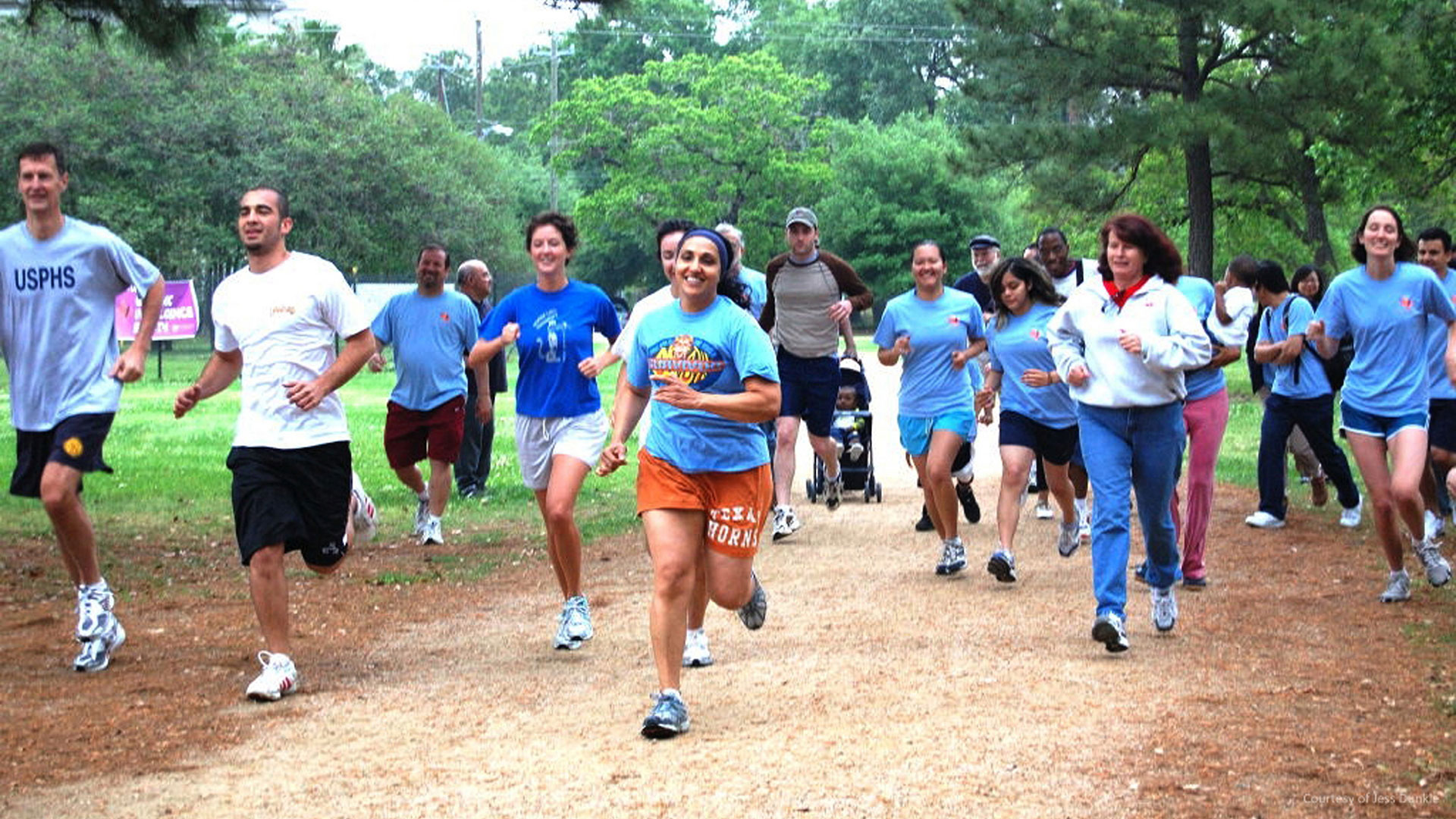 Group of adults jogging toward the camera on a park trail, led by a smiling woman in a blue T-shirt; others follow, including one pushing a stroller, with trees in the background.