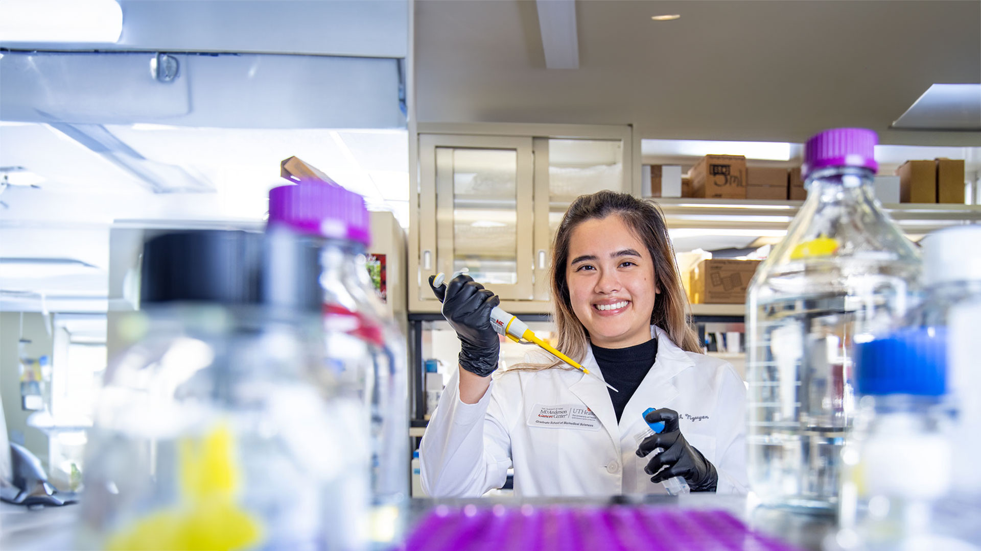 Graduate researcher working in a laboratory with scientific equipment and test bottles in the foreground.